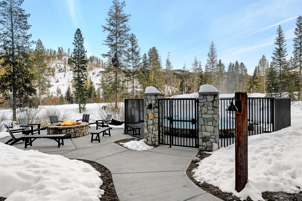 This image depicts a snow-covered outdoor area featuring a cozy fire pit and an inviting hot tub surrounded by pine trees. The scene is set for social gatherings, relaxation, and enjoying the beautiful mountain landscape in all seasons.