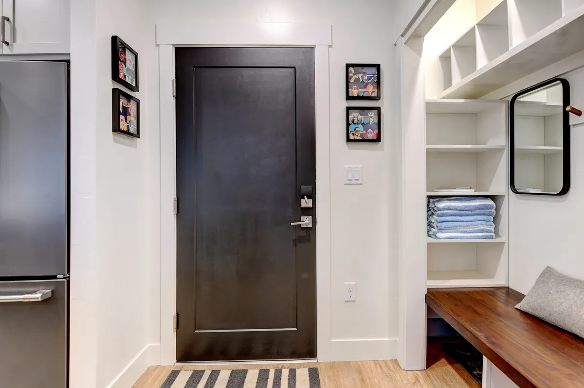 The image shows a welcoming entryway with a black front door, framed photos on the wall, shelves stacked with towels, a mirror, and a wooden bench. The arrangement gives a first impression of the home as inviting and thoughtfully organized for families arriving after a day of fun.