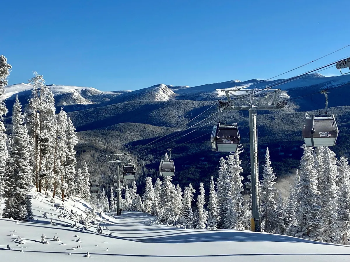 This image features a scenic exterior view showcasing a gondola lift ascending above snow-covered trees with a majestic mountain range in the background. The landscape evokes a sense of adventure and captures the core outdoor activities of skiing, snowboarding, and mountain hiking available at Winter Park Resort.