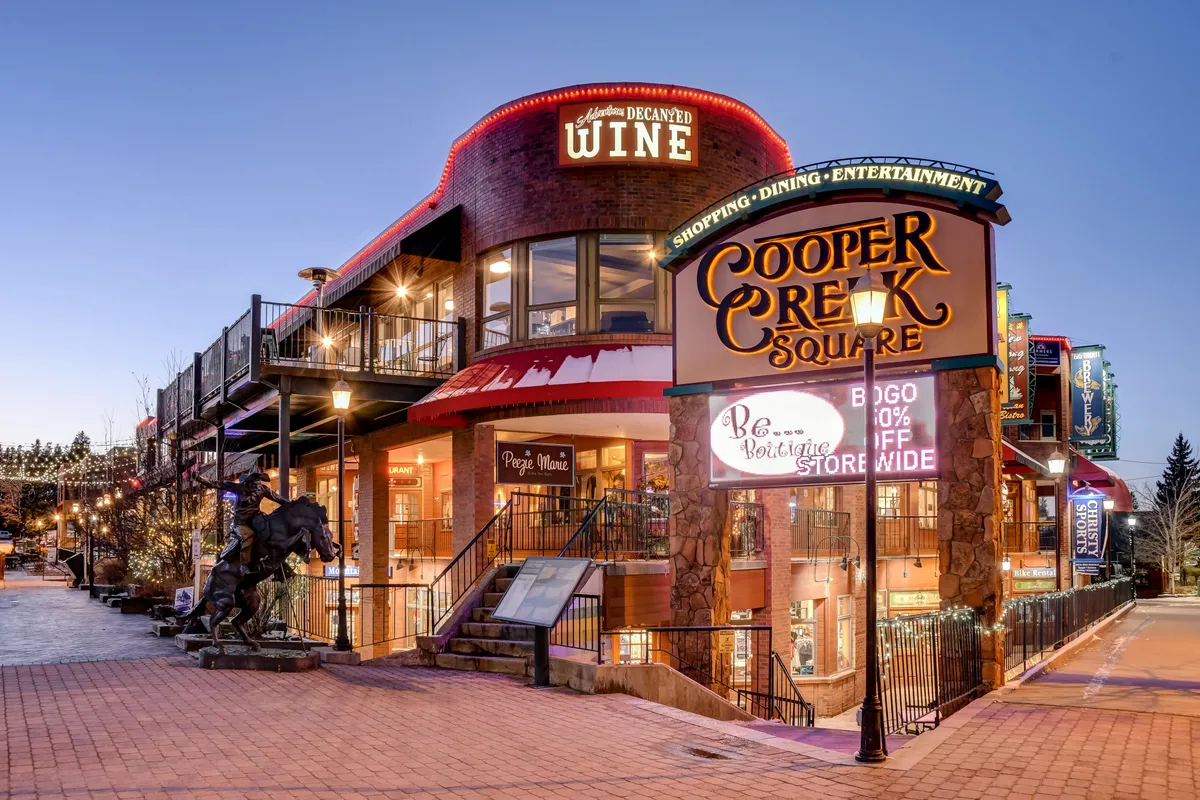 An exterior view of Cooper Creek Square with prominent signage, a wine shop, outdoor dining area, and a striking horse sculpture. The space is lively, hinting at entertainment and social gatherings, and conveys a sense of local culture and charm.