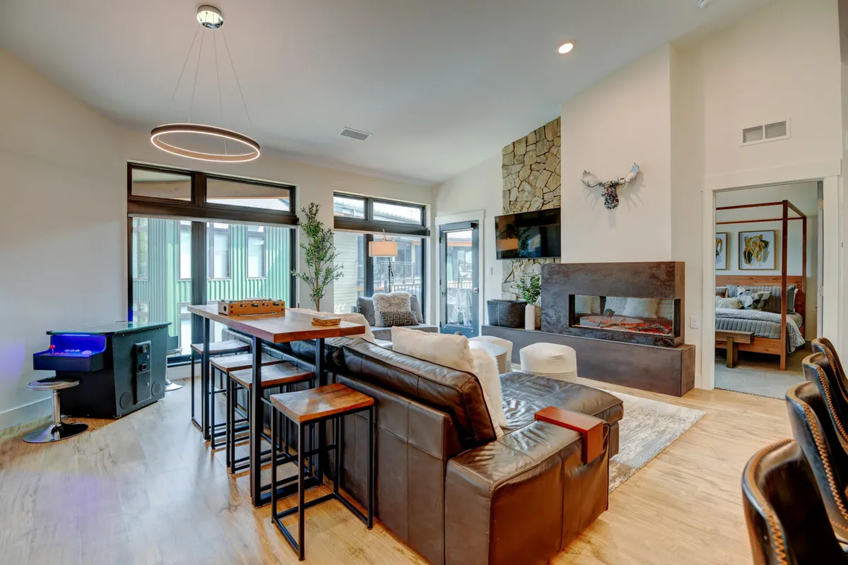 This spacious living area features large windows letting in natural light, a modern fireplace with color-changing features, a stylish dining table, and a comfortable seating area. The space feels bright, open, and perfect for social gatherings, family games, or movie nights.