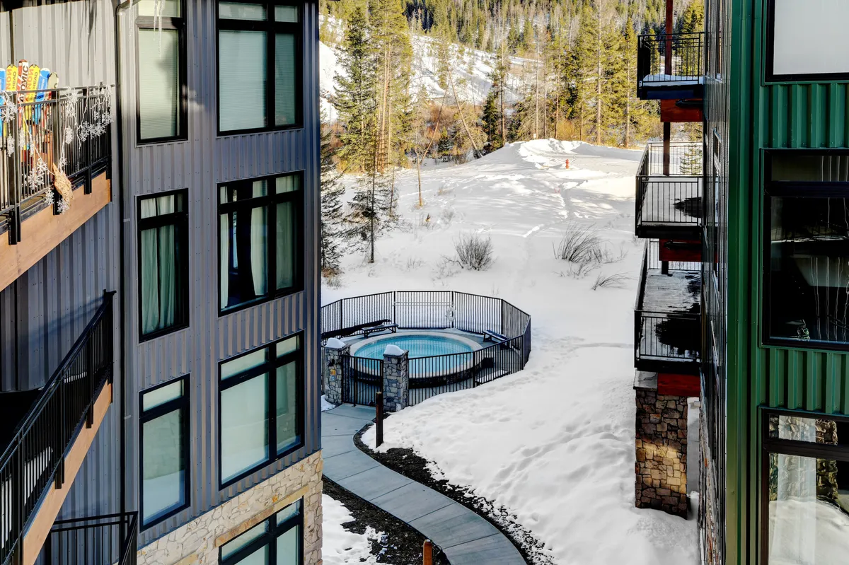 An inviting exterior scene depicting a snow-covered landscape with a cozy hot tub, pathway, surrounding trees, and building facades. The image hints at the relaxation and community atmosphere available after outdoor adventures, and the snowy setting reinforces wintertime activities.
