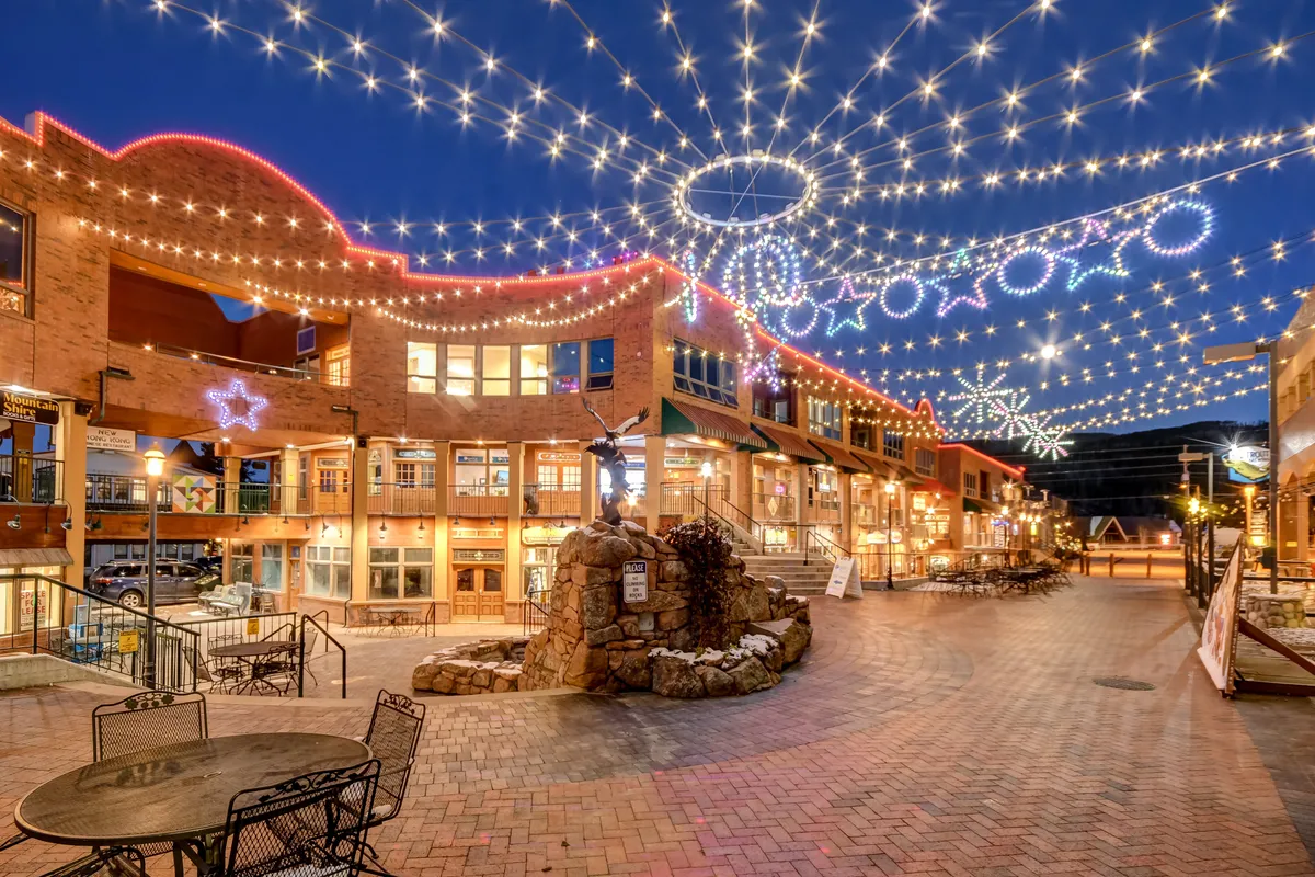 A vibrant outdoor plaza illuminated by string lights, featuring a decorative fountain, bustling shopping complex, and outdoor seating. Shoppers and diners are seen strolling, socializing, and enjoying the festive atmosphere.