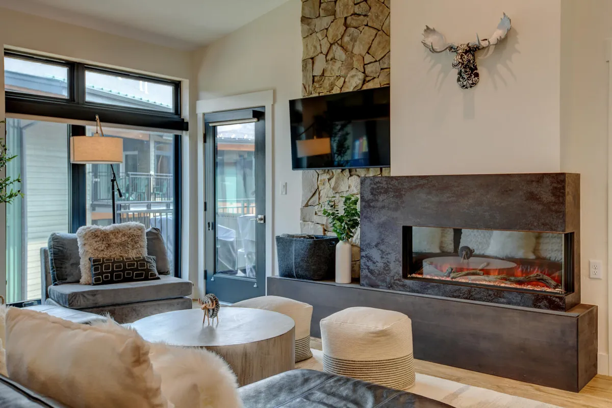 This image showcases a modern fireplace set against a striking stone wall, large windows that bathe the living area in natural light, comfortable seating, and decorative antlers. The arrangement is open and inviting, perfect for both relaxation and social gatherings.