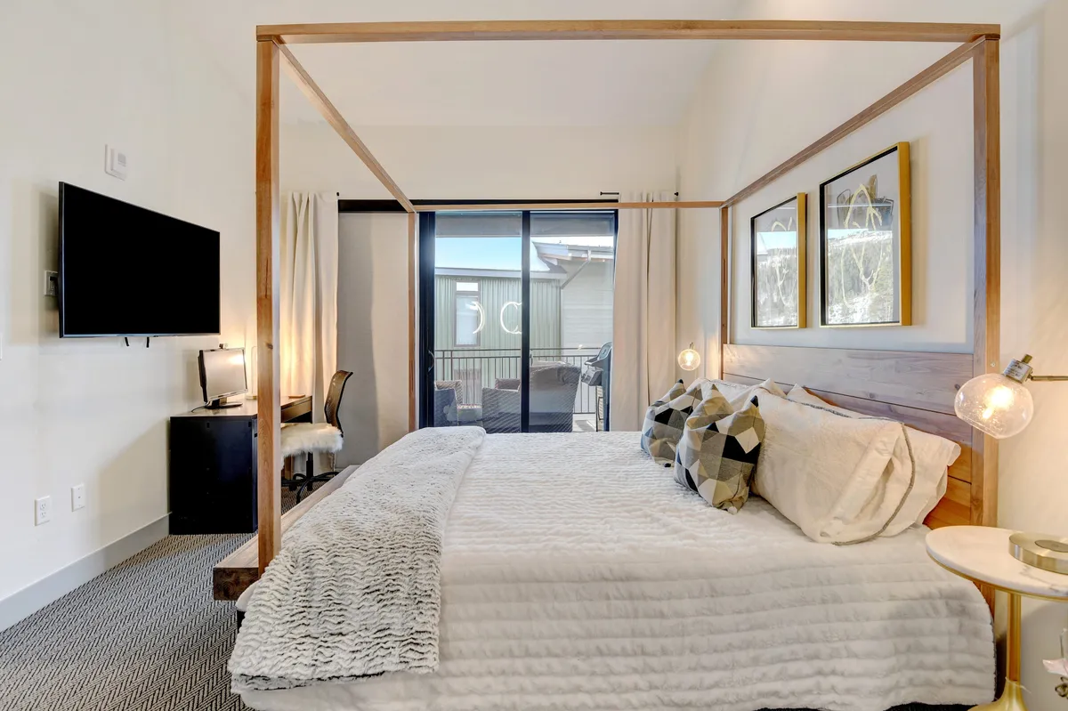 A serene master bedroom featuring a striking four-poster king bed adorned with decorative pillows and luxury linens. Large windows with curtains provide natural light and privacy. A TV is mounted on the wall opposite the bed, and a small desk sits nearby, enhancing both relaxation and functionality.