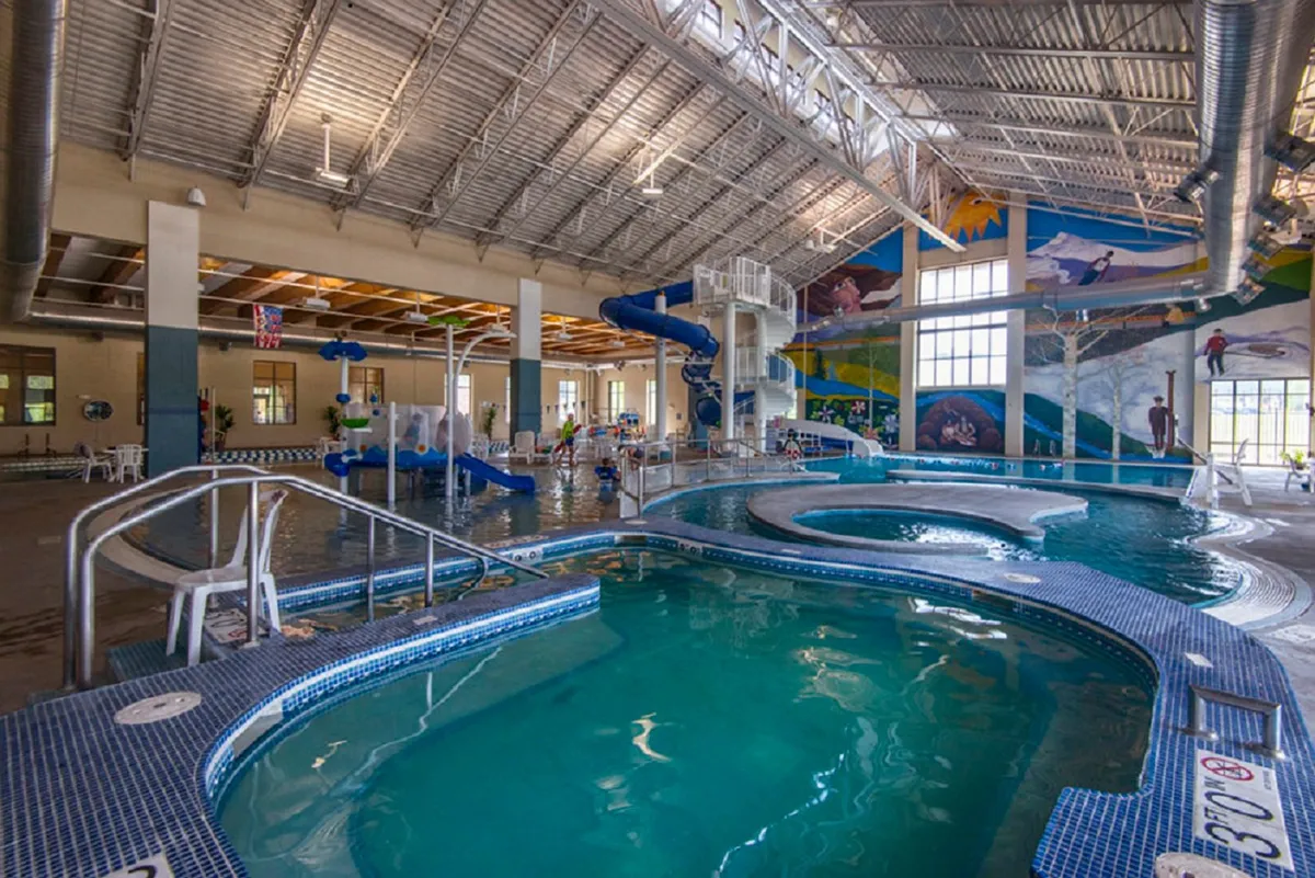 A lively indoor recreational space featuring water slides, swimming pools, interactive water features, and colorful murals. Families and children are seen enjoying aquatic activities and play.