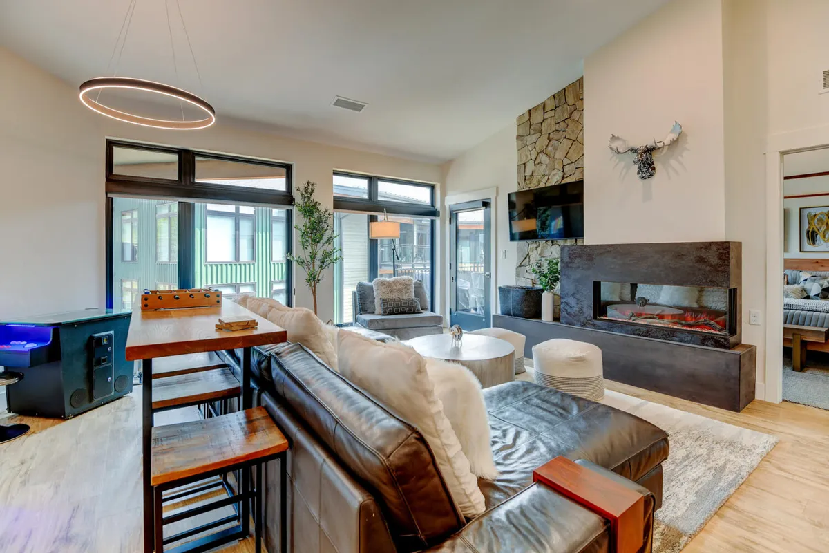 A spacious living room features a stone fireplace, modern chandelier, sectional sofa, and expansive windows offering views of the outdoors. The space is designed for both comfort and elegant entertaining.