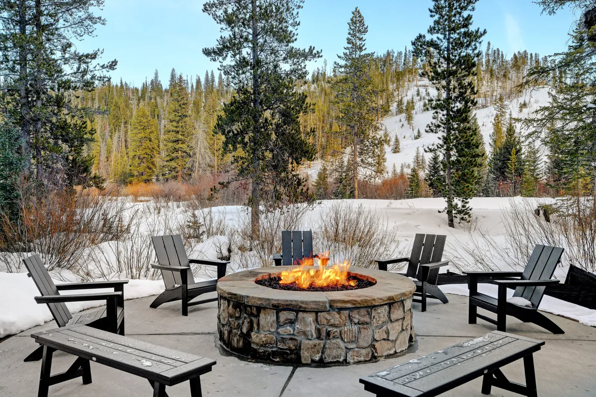 A communal fire pit surrounded by chairs, set on snow-covered ground with tall pines in the background. The scene radiates warmth and togetherness, perfect for gatherings after a day of adventure.