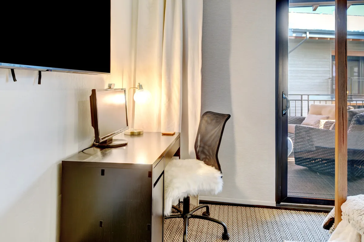 This image shows a dedicated workspace featuring a desk with a computer, a modern chair, and a large window letting in natural light. The setup is complemented by a television and a view of a balcony, creating an inviting and productive atmosphere for remote work.