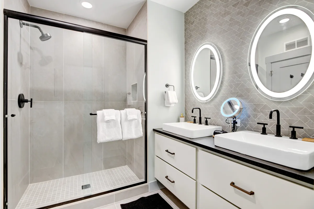 A modern bathroom showcasing a spacious glass walk-in shower, double vanity sinks, and mirrored wall fixtures. The clean lines and gentle lighting create a spa-like ambiance, echoing high-end relaxation and self-care routines.