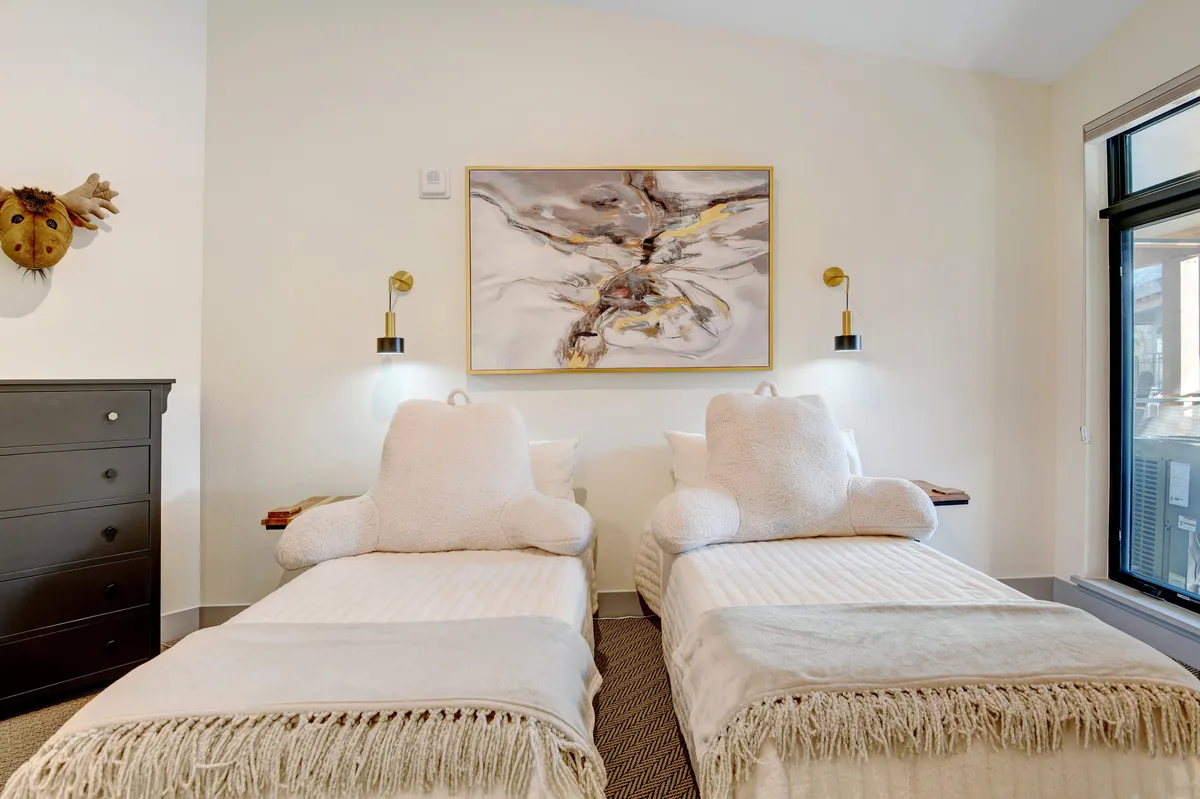 A cozy second bedroom designed for comfort, with twin beds featuring plush headrests, whimsical abstract wall art, and a welcoming side table with a lamp. Natural light spills in, highlighting the inviting atmosphere perfect for kids or friends.